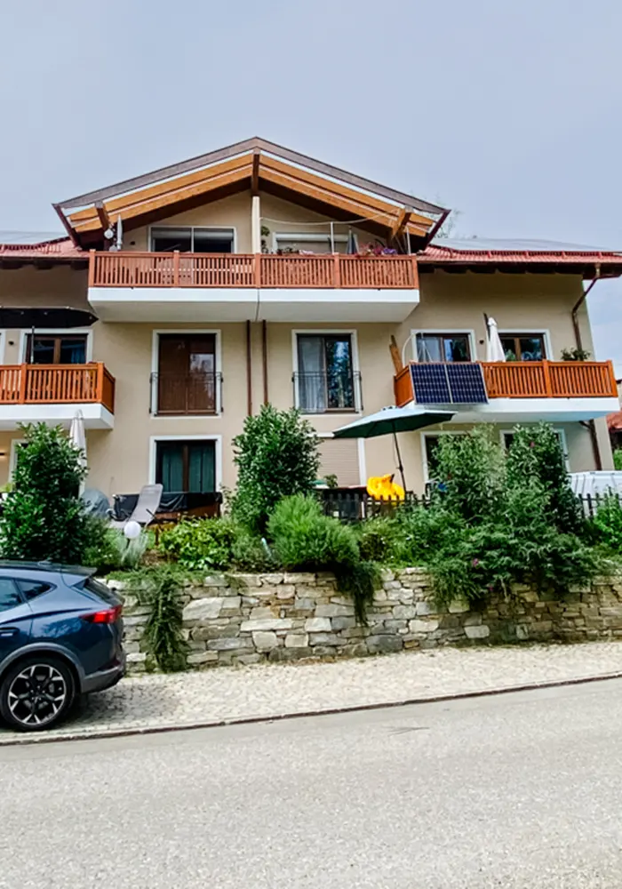 Wohnhaus mit Satteldach, Holzbalkonen und Vorgarten mit Mauer, Pflanzen und Sonnenschirm.