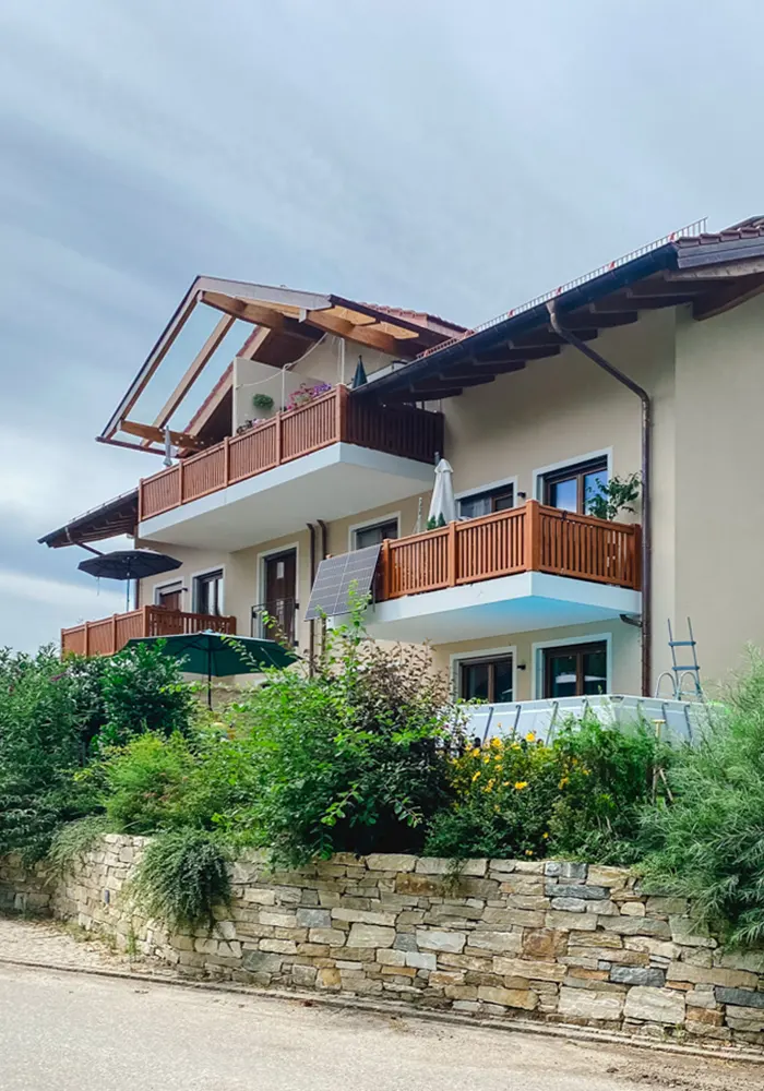 Wohnhaus mit Holz-Balkonen, Solarpanel an der Fassade und dichter Begrünung vor Natursteinmauer.