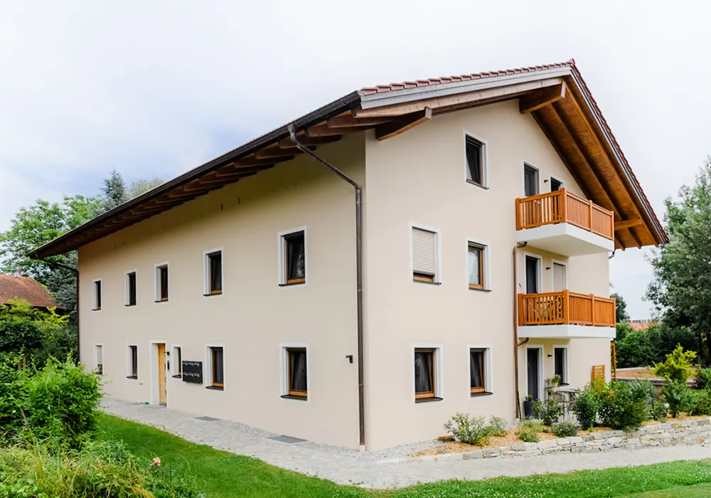 Beiges Mehrfamilienhaus mit Holz-Balkonen, Satteldach und umliegendem Gartenweg.