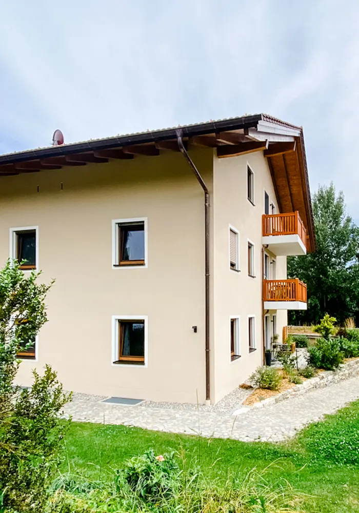 Beiges Wohnhaus mit Holzbalkonen und gepflastertem Weg, umgeben von Rasen und Pflanzen.