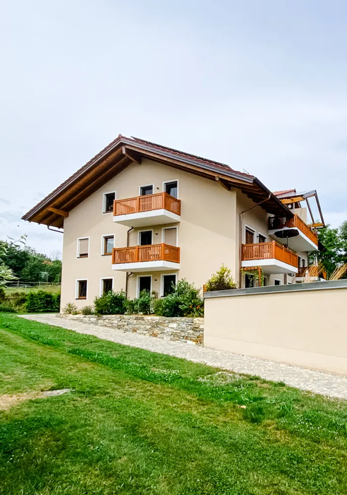 Beiges Mehrfamilienhaus mit roten Holzbalkonen, Gartenweg und Grünfläche im Vordergrund.