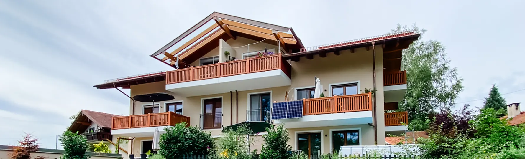 Mehrfamilienhaus mit Holz-Balkonen, Glasdach, Solarpanelen und üppiger Bepflanzung im Vorgarten.