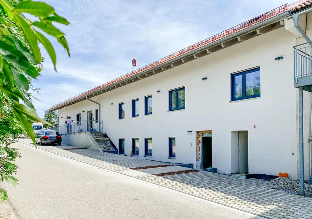 Langes, weißes Gebäude mit blauen Fenstern an einer leicht abschüssigen Straße, Autos parken davor.