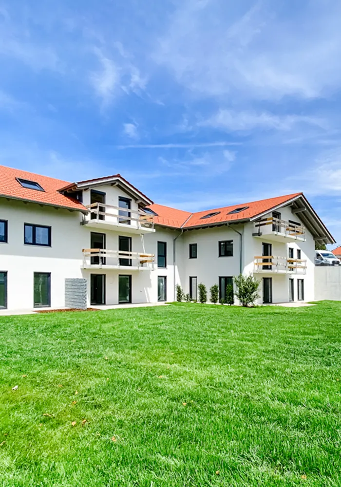 Mehrfamilienhaus mit rotem Satteldach, weißen Balkonen und begrünter Gartenanlage mit Baum und Sitzbank.