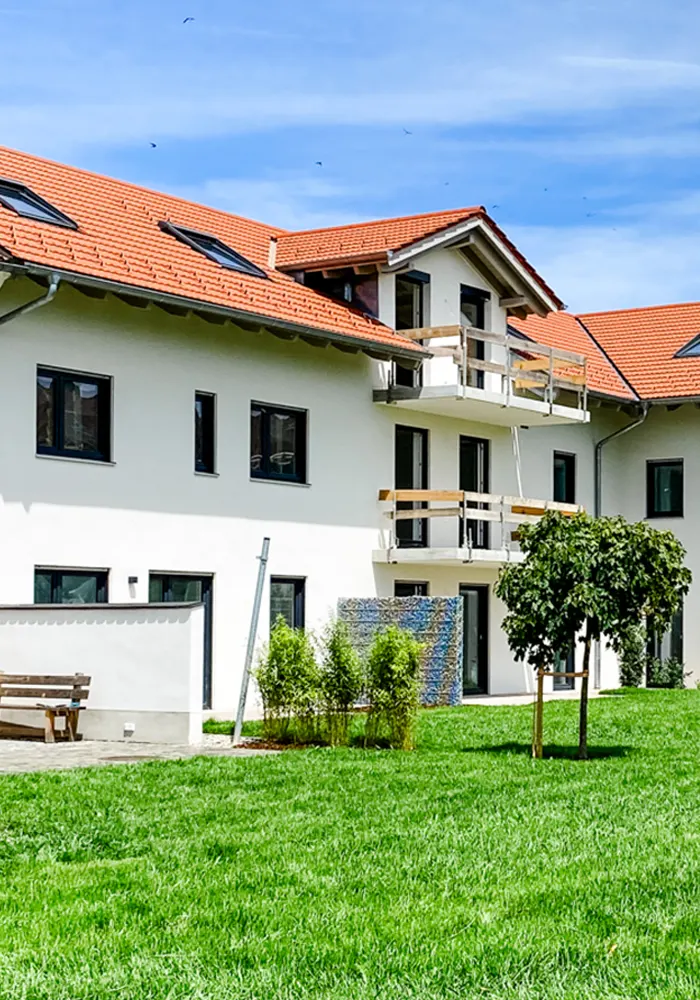 Mehrfamilienhaus mit rotem Satteldach, weißen Balkonen und begrünter Gartenanlage mit Baum und Sitzbank.