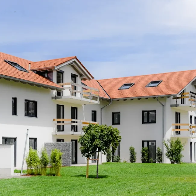 Mehrfamilienhaus mit rotem Satteldach, weißen Balkonen und begrünter Gartenanlage mit Baum und Sitzbank.
