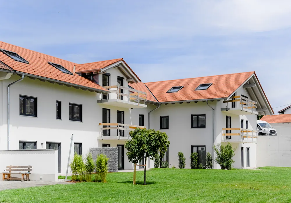 Mehrfamilienhaus mit rotem Satteldach, weißen Balkonen und begrünter Gartenanlage mit Baum und Sitzbank.