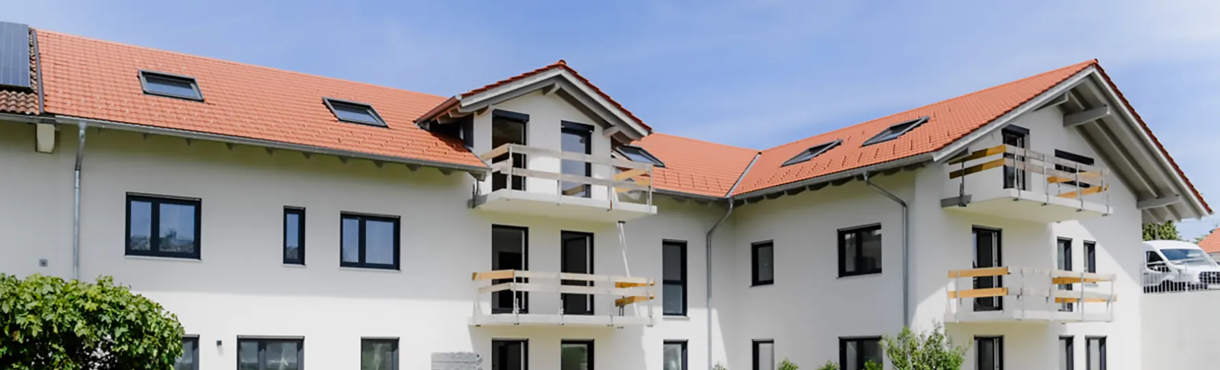 Mehrfamilienhaus mit rotem Satteldach, weißen Balkonen und begrünter Gartenanlage mit Baum und Sitzbank.