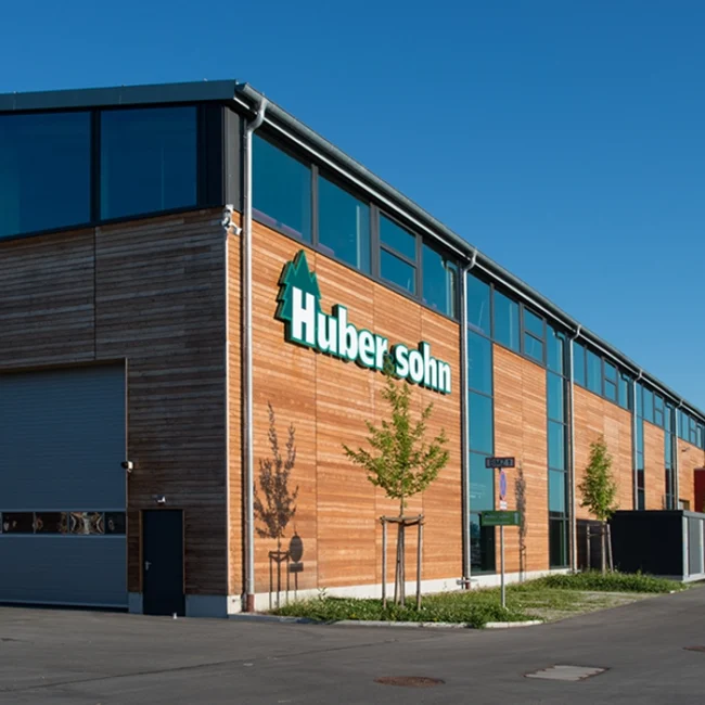 Modernes Industriegebäude mit Holzfassade und Aufschrift ‚Huber & Sohn‘ unter blauem Himmel.