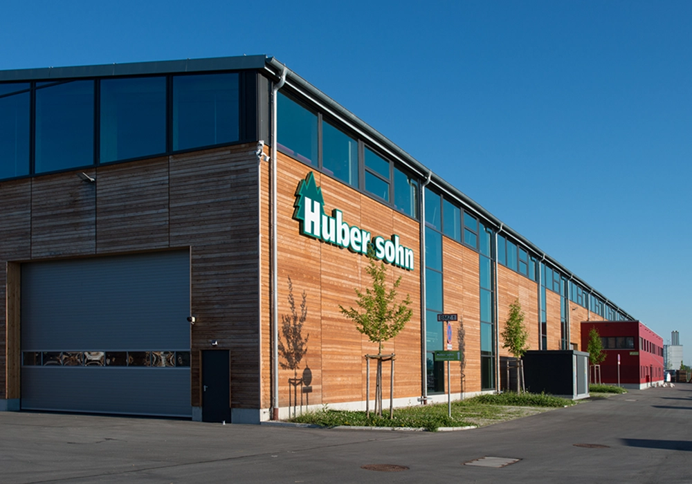 Modernes Industriegebäude mit Holzfassade und Aufschrift ‚Huber & Sohn‘ unter blauem Himmel.