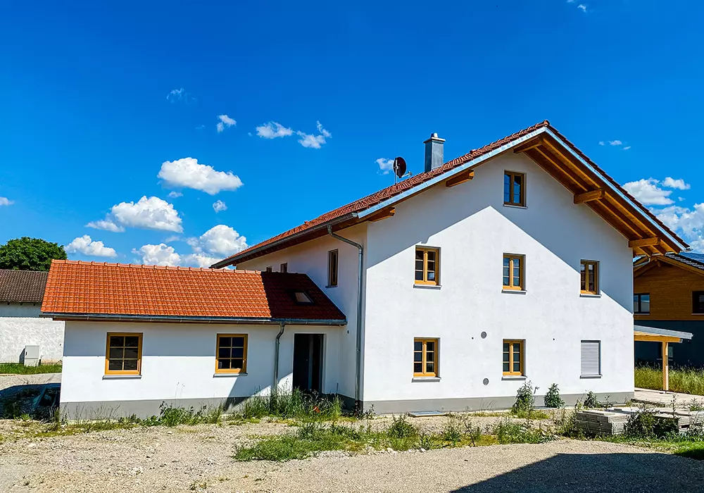 Weißes Wohnhaus mit rotem Ziegeldach und Holzfenstern, daneben Anbau und Naturgarten unter blauem Himmel.