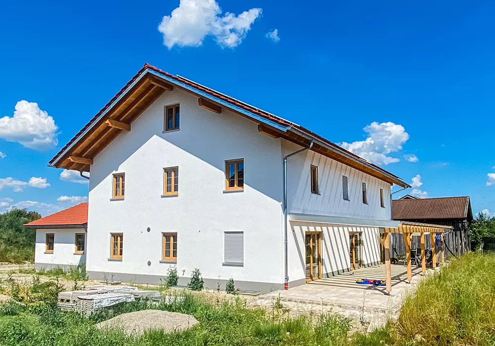 Modernisiertes Landhaus mit weißer Fassade, rotem Ziegeldach, Holzfenstern und Pergola auf Terrasse, umgeben von Wiese.