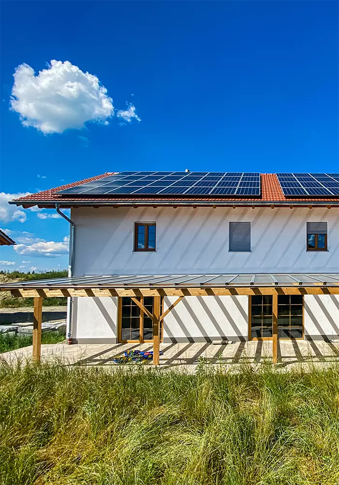 Modernes Wohnhaus mit Solaranlage auf dem Dach und Holzpergola auf Terrasse, umgeben von hoher Wiese.