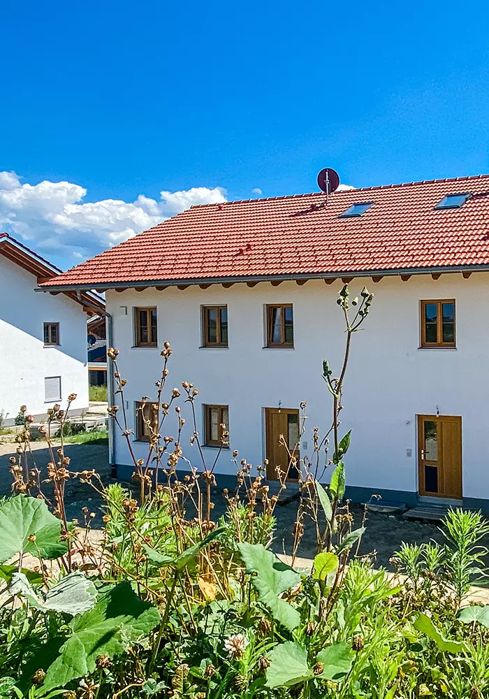 Weißes Wohnhaus mit rotem Ziegeldach und Holzfenstern, dahinter blauer Himmel, vorn hohe Wildpflanzen.