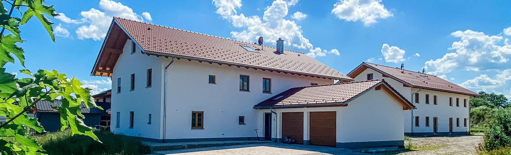 Zwei moderne Wohnhäuser mit rotem Satteldach, Garage im Vordergrund, unter blauem Himmel mit wenigen Wolken.