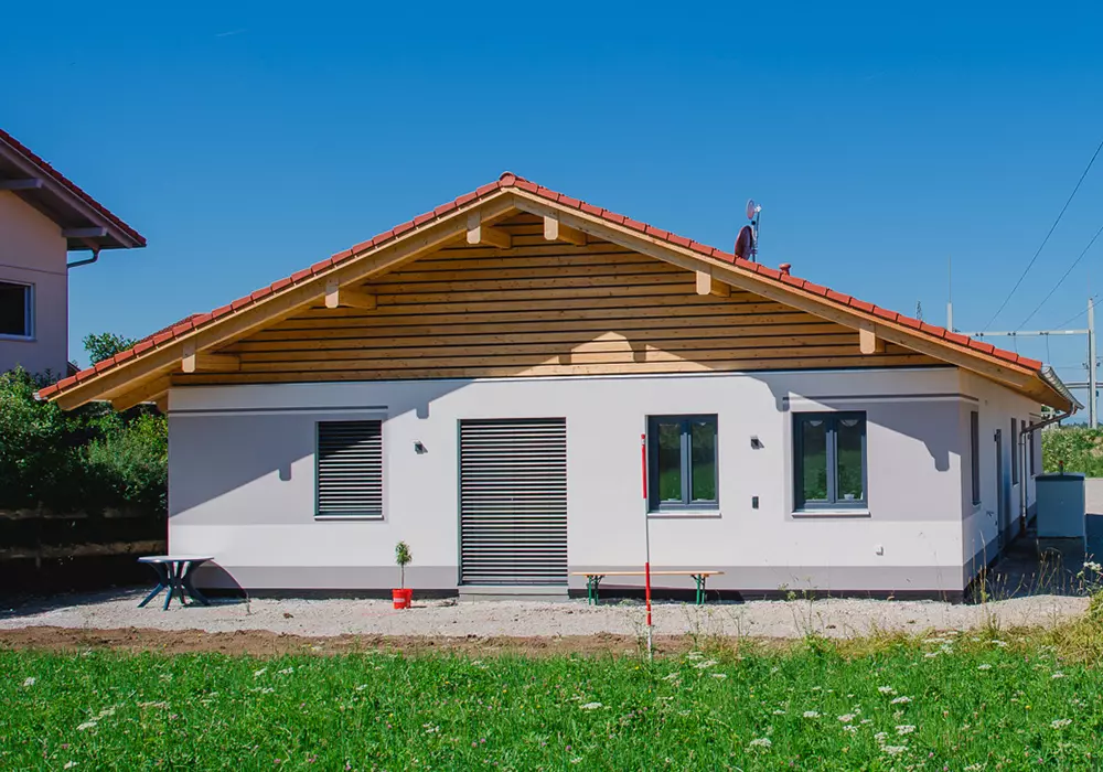 Erdgeschossiger Neubau mit Satteldach, Holzfassade im Giebel, Rollläden und kleiner Sitzgruppe vor dem Haus.