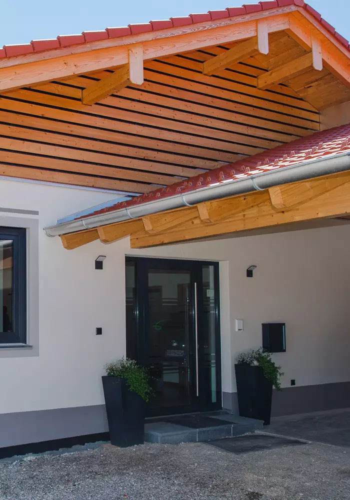 Moderner Flachbau mit rotem Ziegeldach, Wärmepumpe, Carport und Glas­eingang unter Holzvordach bei sonnigem Wetter.