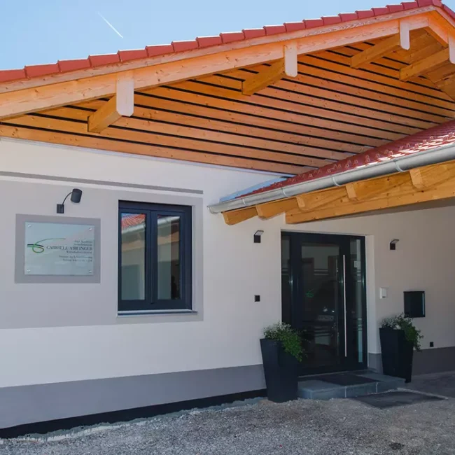 Eingangsbereich mit Vordach aus Holz, Glasfront, Namensschild und Auto unter Carport vor einem modernen Flachbau.