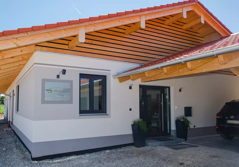 Eingangsbereich mit Vordach aus Holz, Glasfront, Namensschild und Auto unter Carport vor einem modernen Flachbau.