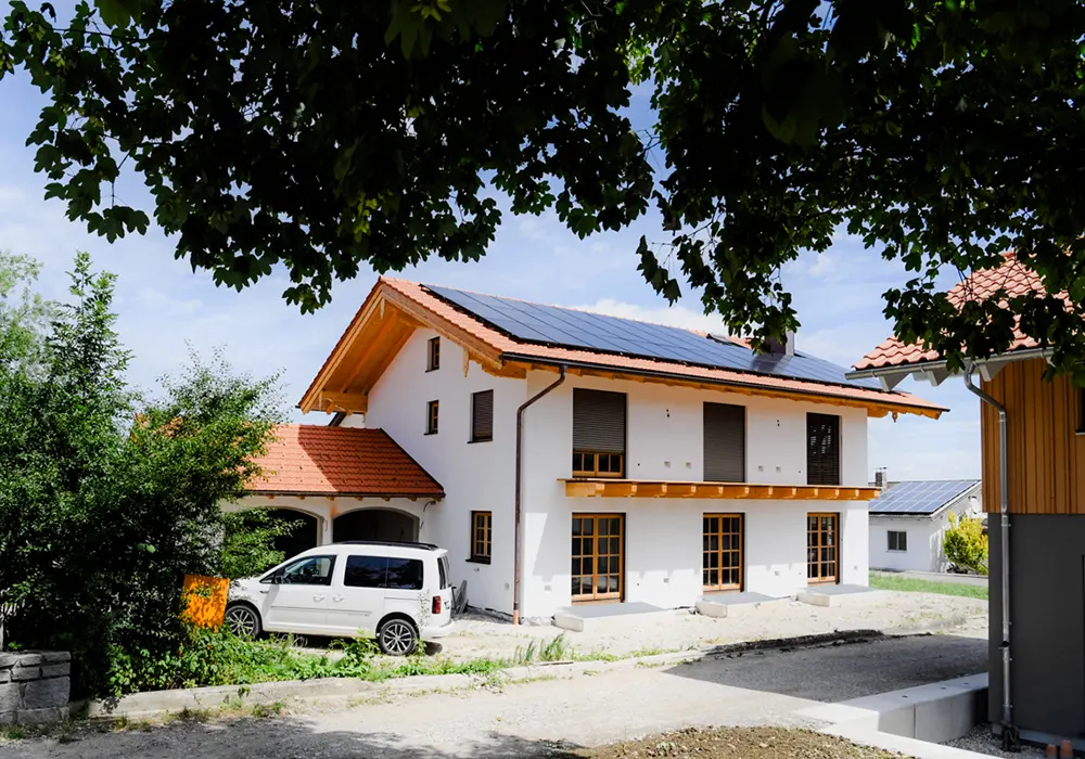 Modernes Haus mit Holzdetails und Solarpanels, weißer Van davor, Bäume rahmen das Bild.