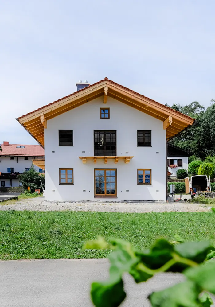 Frontansicht eines weißen Hauses mit Holzgiebel, geschlossenen Rollläden und Garten im Vordergrund.