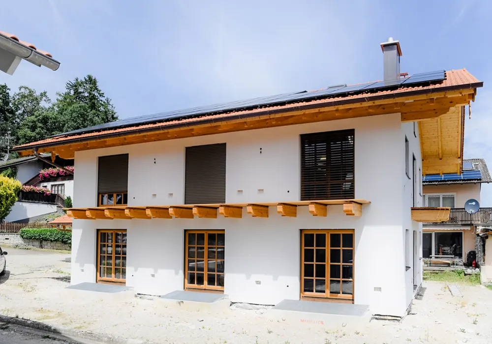 Zweistöckiges Haus mit Solaranlage, Holzfenstern, Balkenüberstand und geschlossenen Rollläden, daneben Nachbarhäuser.