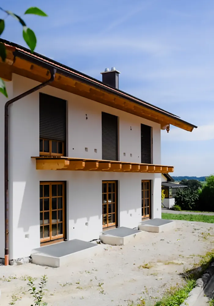 Rückseite eines Hauses mit Holzfenstern, geschlossenen Rollläden und kleinen Betonterrassen auf unbepflanztem Grundstück.