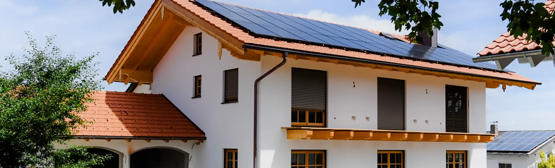 Haus mit rotem Ziegeldach, geschlossenen Rollläden und großer Solaranlage auf dem Dach, daneben Baum und Nachbarhaus.