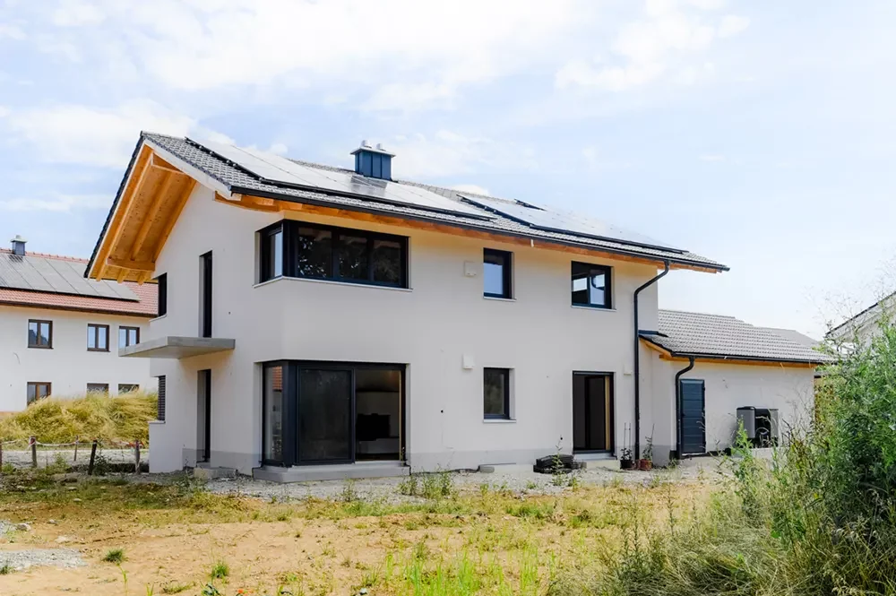 Rückansicht eines modernen Hauses mit Solaranlage auf dem Dach, dunklen Fenstern und unbepflanztem Gartenbereich.