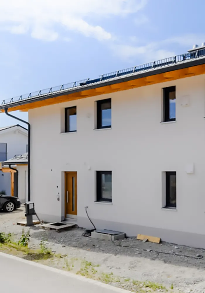 Vorderansicht eines neuen Einfamilienhauses mit dunklen Fenstern, Holzvordach und unbefestigtem Eingangsbereich.