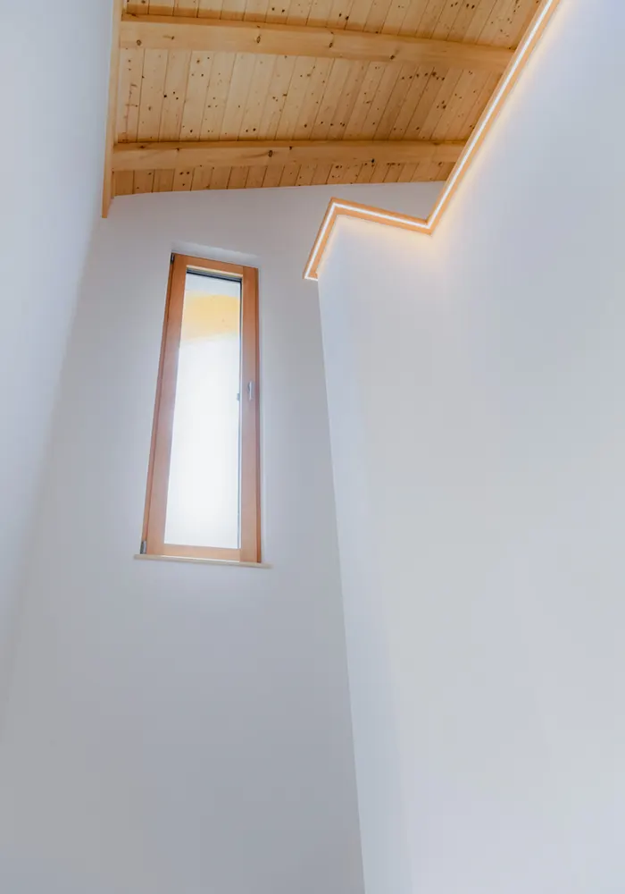 Heller Innenraum mit schrägem Holzdeckengebälk, hohem Fenster und indirekter LED-Beleuchtung an der Wand.