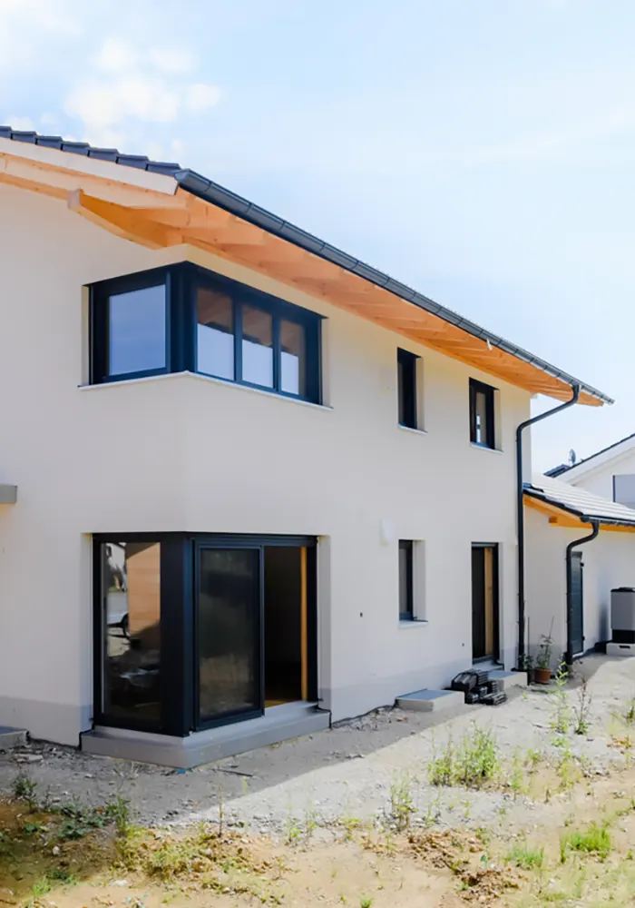 Neubau eines hellen Einfamilienhauses mit dunklen Fensterrahmen und unfertigem Gartenbereich im Vordergrund.