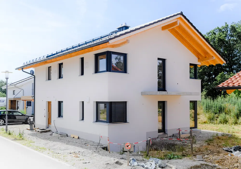 Eckansicht eines neuen, hellen Einfamilienhauses mit dunklen Fenstern, Holzgiebel und unfertigem Vorgarten.