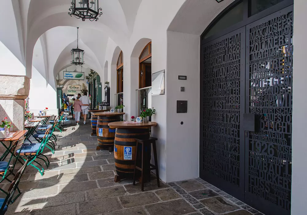 Überdachter Arkadengang mit Sitzplätzen, Stehtischen aus Holzfässern und kunstvoller Metalltür mit Schriftprägung.