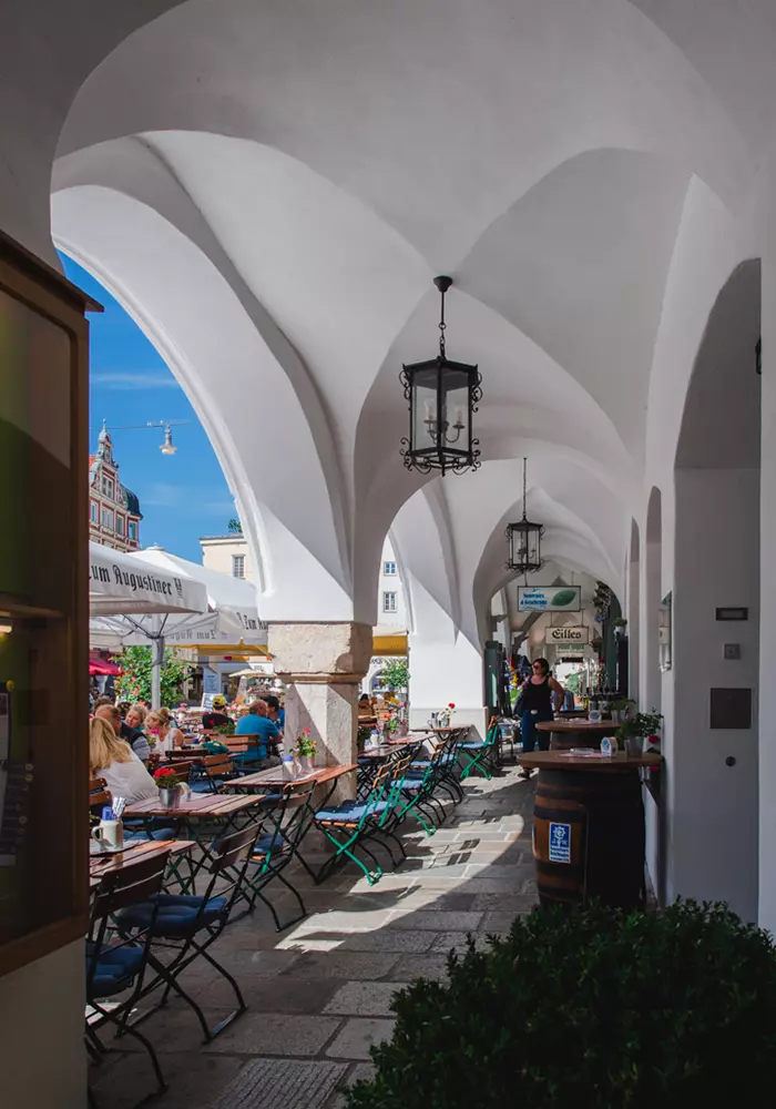 Gewölbter Arkadengang mit Hängelampen und Cafébestuhlung, daneben belebter Außenbereich bei sonnigem Wetter.