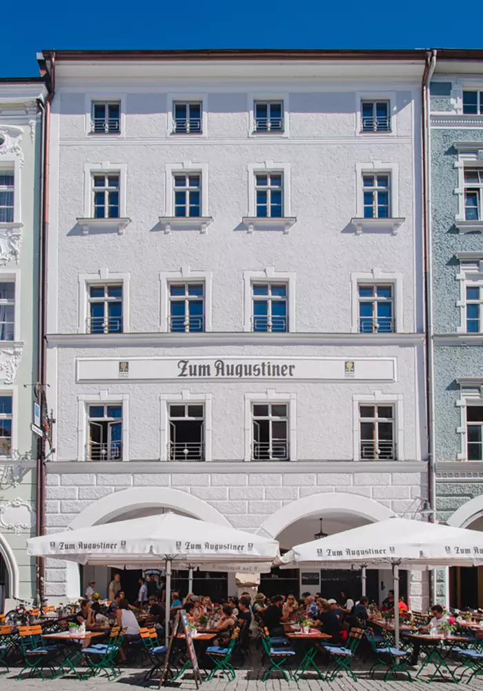 Historisches Gebäudeensemble mit Restaurant „Zum Augustiner“, weißer Fassadenputz, Rundbogenarkaden und Außenbestuhlung auf Kopfsteinpflaster unter Sonnenschirmen.
