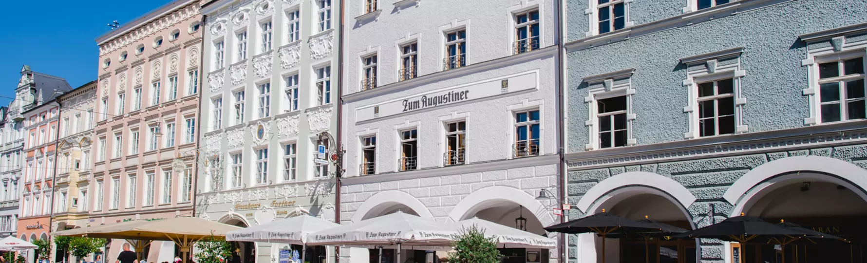 pauker-bau-gmbh-referenzen-putz-sanierung-fassade-wohn-geschaeftshaus-rosenheim-header Historisches Gebäudeensemble mit Restaurant „Zum Augustiner“, weißer Fassadenputz, Rundbogenarkaden und Außenbestuhlung auf Kopfsteinpflaster unter Sonnenschirmen.