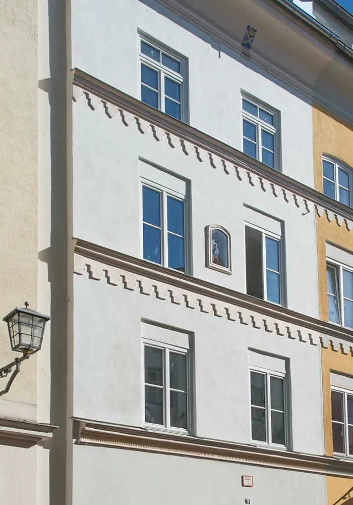 Weiße Altbaufassade mit Zierleiste, weißen Fenstern und religiöser Wandnische, daneben gelb gestrichenes Nachbarhaus.