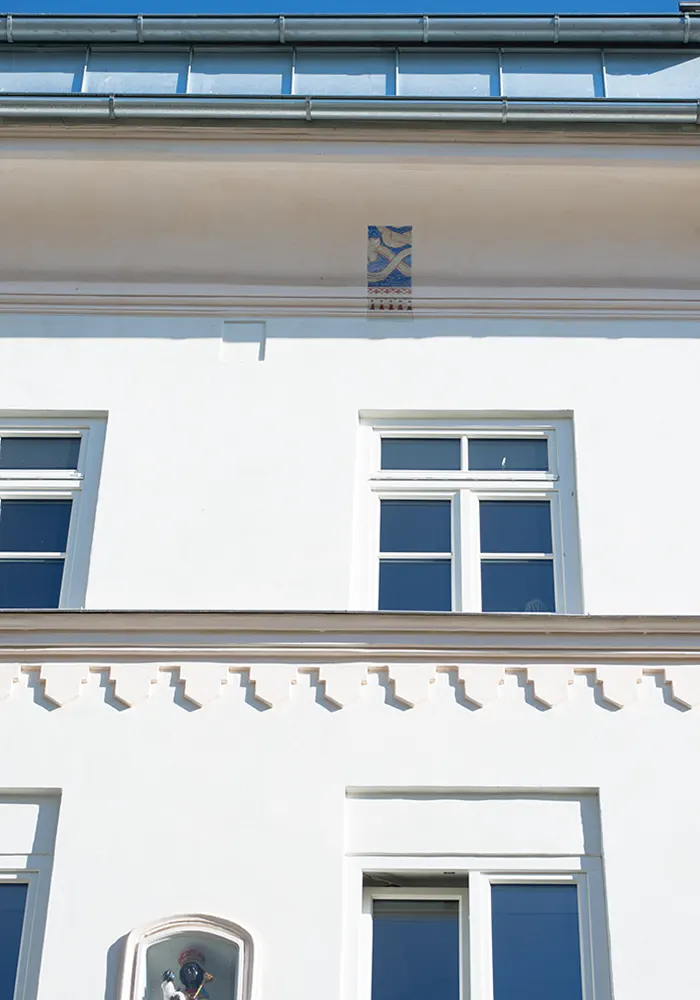 Ausschnitt einer weißen Hausfassade mit Zierleiste, weißen Fenstern und bemalter Wandnische unterhalb des Dachs.