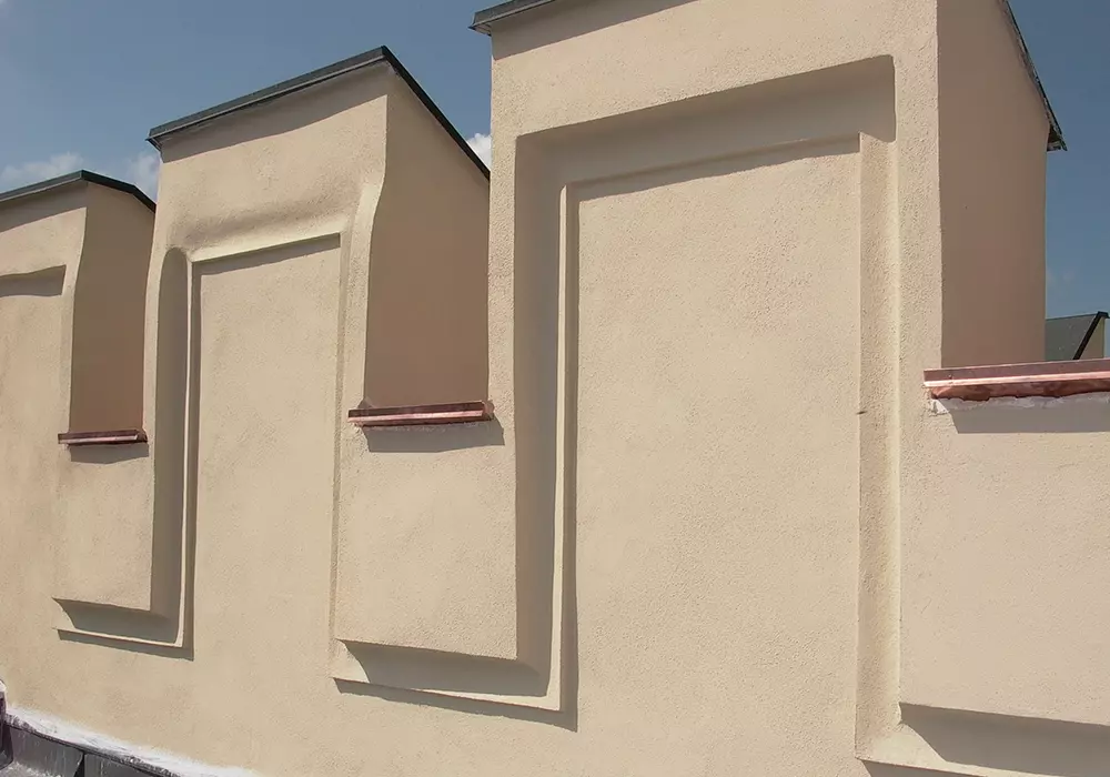 Neu verputzte, beige Wand mit dekorativen Nischen und Kupferabdeckungen unter blauem Himmel.