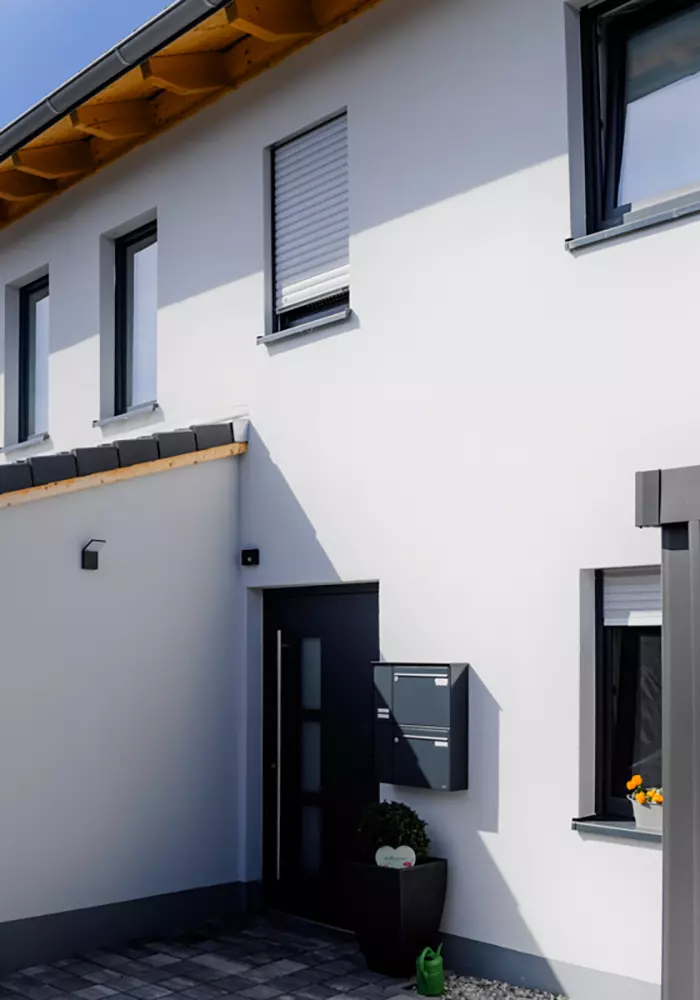 Hauseingang mit dunkler Tür, Briefkasten, Pflanzkübel und Fenster mit Blumen unter hellem Putz und Satteldach.