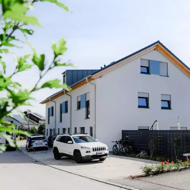 Modernes weißes Wohnhaus mit Satteldach, parkenden Autos und Fahrrädern in ruhiger Wohnstraße bei Sonnenschein.