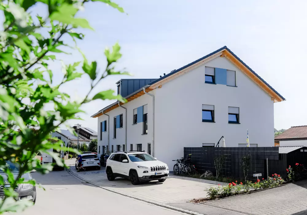 Modernes weißes Wohnhaus mit Satteldach, parkenden Autos und Fahrrädern in ruhiger Wohnstraße bei Sonnenschein.