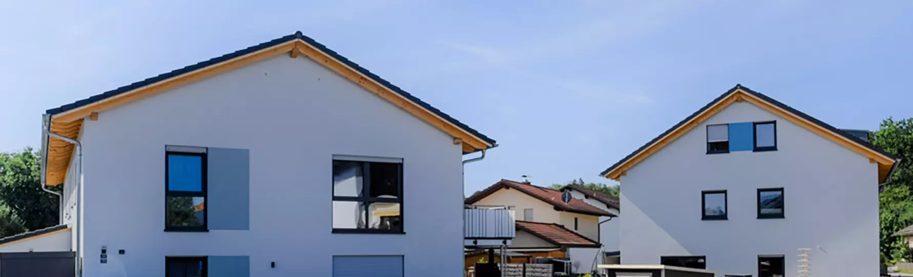 Zwei moderne Einfamilienhäuser mit Satteldach und blauen Fensterläden bei klarem Himmel in ruhiger Wohngegend.