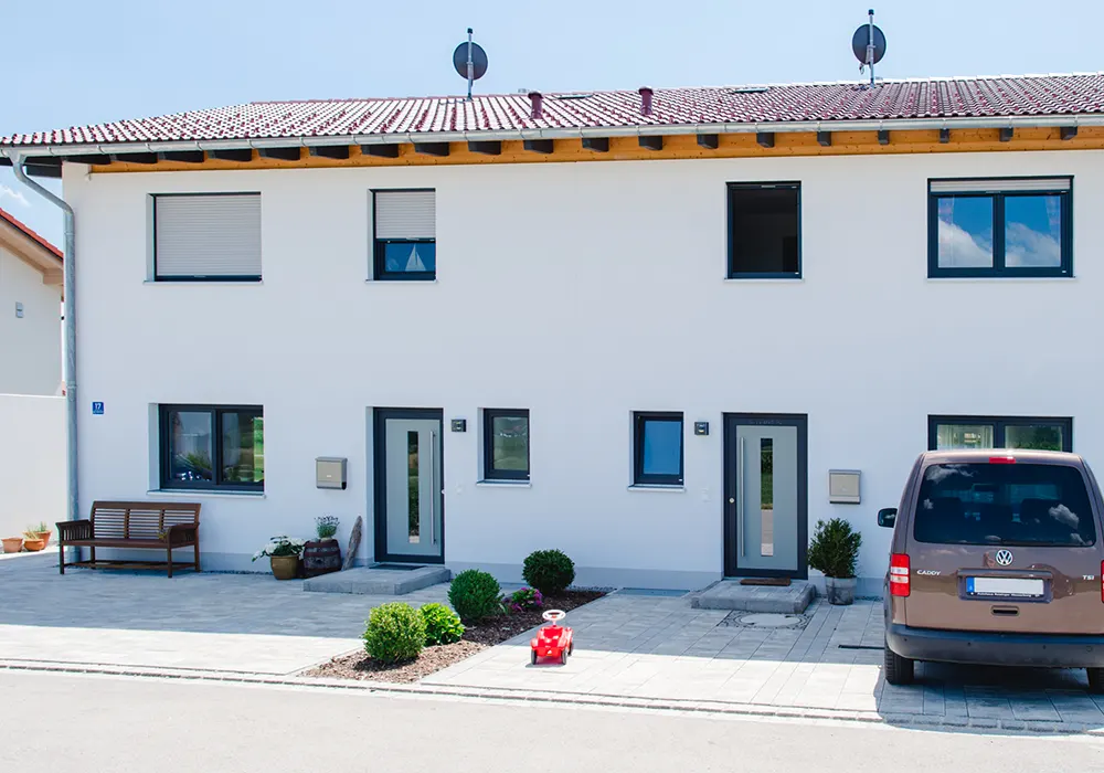 Vorderansicht eines modernen Doppelhauses mit rotem Dach, Vorgarten, geparktem Auto und rotem Kinderrutscher.