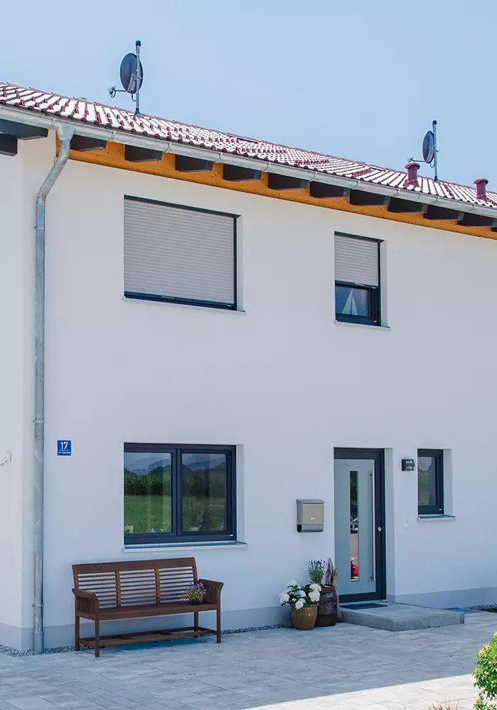 Eingangsbereich eines weißen Hauses mit Bank, Blumentöpfen und geschlossenen Rollläden.