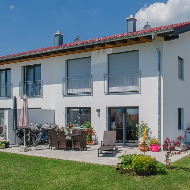 Modernes weißes Einfamilienhaus mit rotem Satteldach, mehreren Balkontüren und kleinen Balkonen im Obergeschoss; davor eine möblierte Terrasse mit Stühlen, Tisch, Sonnenliegen und Blumen, daneben einige Fahrräder.