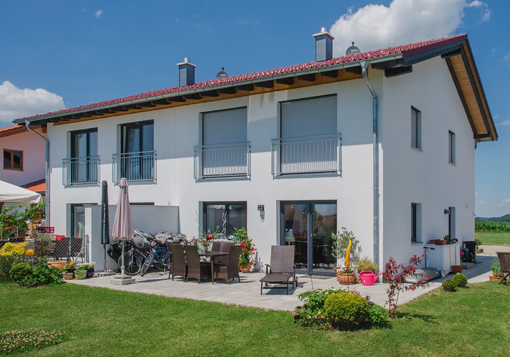 Modernes weißes Einfamilienhaus mit rotem Satteldach, mehreren Balkontüren und kleinen Balkonen im Obergeschoss; davor eine möblierte Terrasse mit Stühlen, Tisch, Sonnenliegen und Blumen, daneben einige Fahrräder.