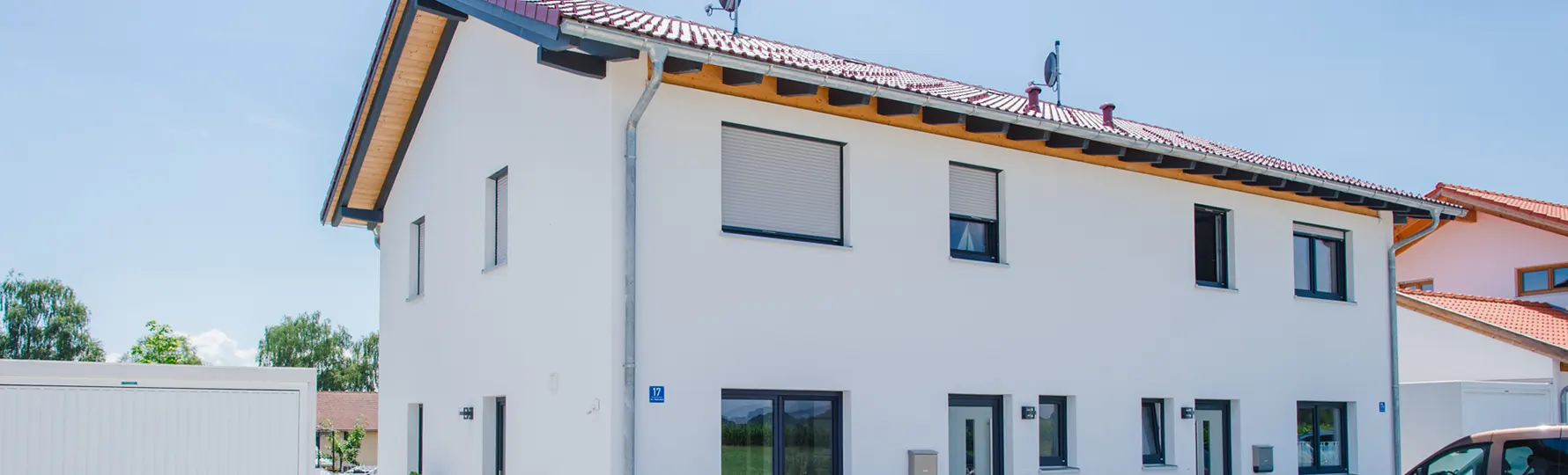 Modernes, weißes Zweifamilienhaus mit roten Ziegeldach und Rollläden, daneben Garage und geparktes Auto.
