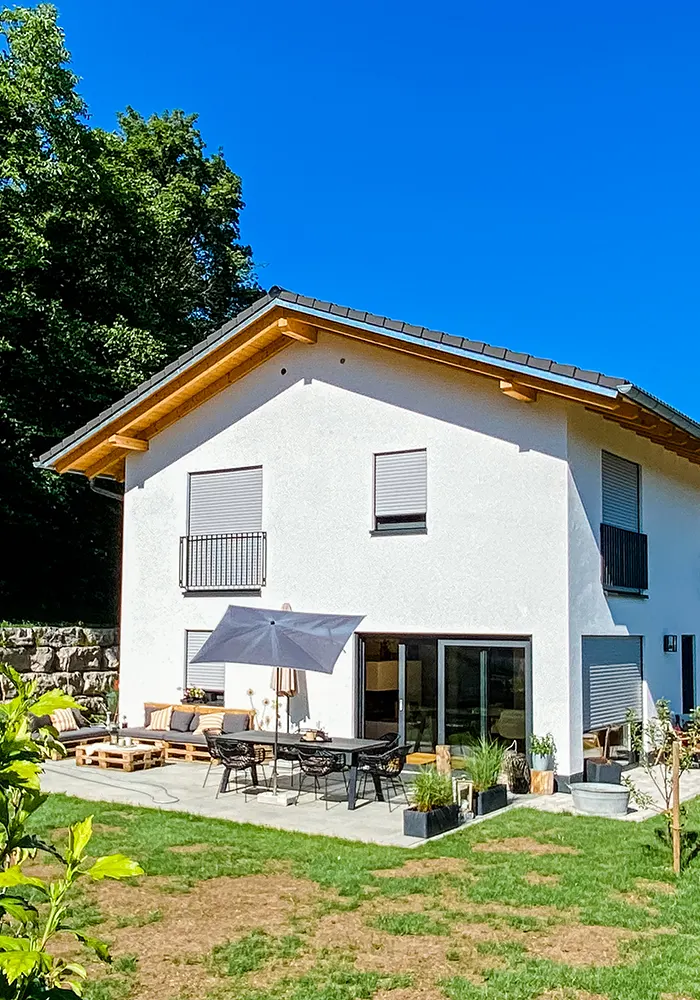 Zweigeschossiges Haus mit Terrasse, Gartenmöbeln und Sonnenschirm, davor Rasenfläche mit braunen Stellen.